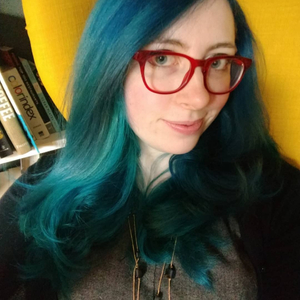 A photo of Ashley Kolodziej. She has blue hair and wears a bright yellow cardigan and red glasses.